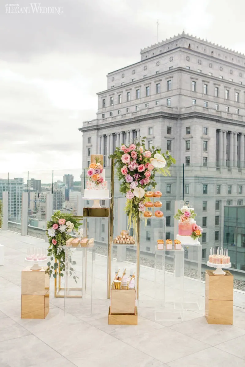 The image features a stunning rooftop wedding cake and dessert display by Pâtisserie Montebello, showcasing their expertise in the Wedding Cakes category. Centered around an exquisite tiered cake adorned with pastel flowers, the arrangement includes a variety of elegant pastries and cupcakes. The desserts are artfully placed on modern, gold and glass stands, enhancing the luxurious theme. The backdrop includes an urban cityscape with a prominent building, adding a sophisticated touch to the setting. This visually captivating setup is ideal for a chic and stylish wedding celebration in Canada.
