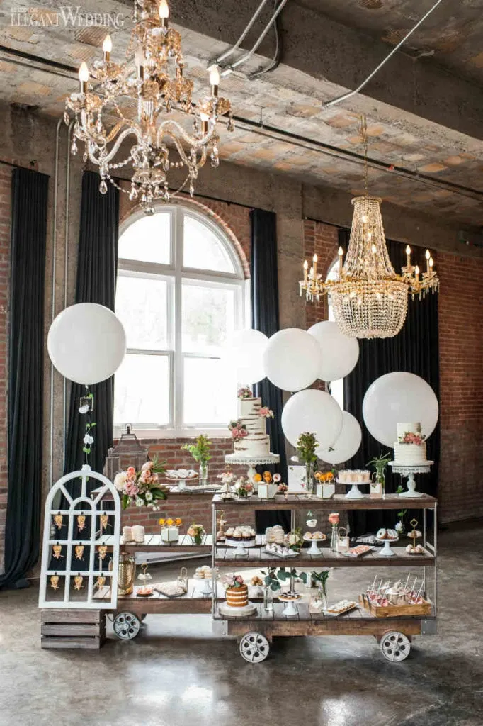 This image features an elegant display by Pâtisserie Montebello, under the wedding cakes category, showcasing a selection of luxurious confections perfect for weddings. The setting is a chic industrial venue with exposed brick walls and large arched windows, illuminated by ornate chandeliers. The centerpiece includes a tiered wedding cake adorned with delicate floral decorations. Surrounding the main cake are various pastries and desserts, artistically arranged, highlighting the vendor's expertise in wedding cakes and desserts. White balloons add a festive touch, complementing the sophisticated atmosphere ideal for an upscale wedding reception.