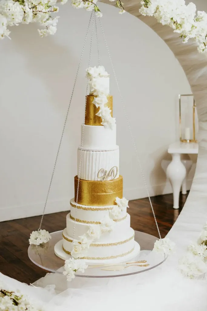 This image showcases an elegant wedding cake created by Pâtisserie Montebello, listed under the Wedding Cakes category. The cake is a multi-tiered masterpiece with alternating layers of shimmering gold and pristine white fondant. It features intricate floral decorations cascading down the tiers, adding a touch of sophistication. Suspended within a circular floral frame, the cake epitomizes luxury and artistry, making it an exquisite choice for a wedding celebration. The initials 'C & D' are elegantly embossed on a gold layer, personalizing the design for the couple. This presentation reflects the high-quality craftsmanship offered by Pâtisserie Montebello for weddings in Canada.