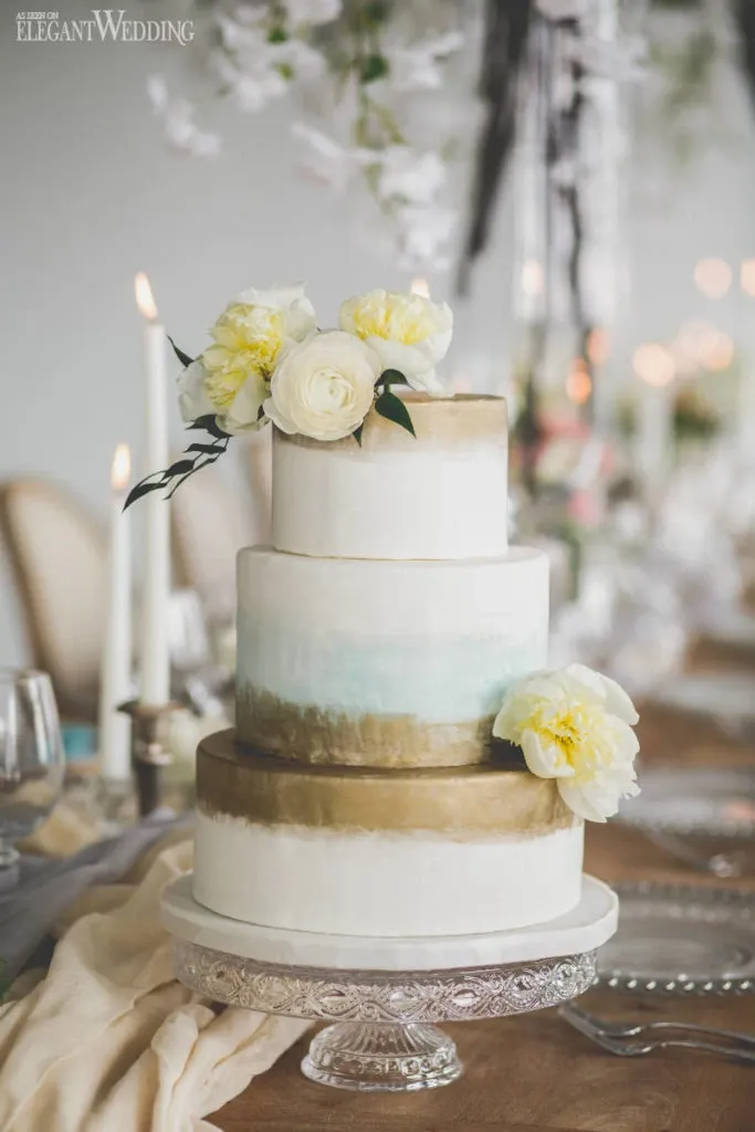 A three-tiered wedding cake by Pâtisserie Montebello, featured in the Wedding Cakes category of Mariage Elegant, Canada. The cake is elegantly decorated with a soft gradient of white, light blue, and gold, creating a sophisticated ombre effect. Each tier is adorned with delicate white and pale yellow peonies, enhancing the cake's luxurious appearance. It is placed on a decorative silver cake stand. In the background, a beautifully set wedding table is visible with tall white candles, elegant glassware, and floral arrangements, contributing to an overall romantic and elegant wedding setting.