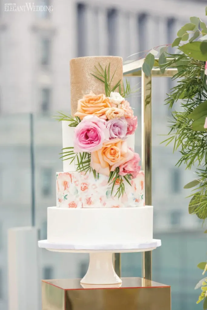 This image showcases a stunning wedding cake by Pâtisserie Montebello, a vendor featured in the wedding category of Mariage Elegant, Canada. The cake is elegantly displayed on a white pedestal with a sleek, modern design. It comprises four tiers, each with distinct styling. The top tier is textured with a shimmering gold finish. Below it, the second tier features a bouquet of pastel-colored roses in pink, peach, and lavender, accented with sprigs of greenery. The third tier has a watercolor floral pattern, adding a touch of artistic flair. The bottom tier is a smooth, classic white. The backdrop of a cityscape enhances the sophisticated ambiance, ideal for a contemporary wedding setting.