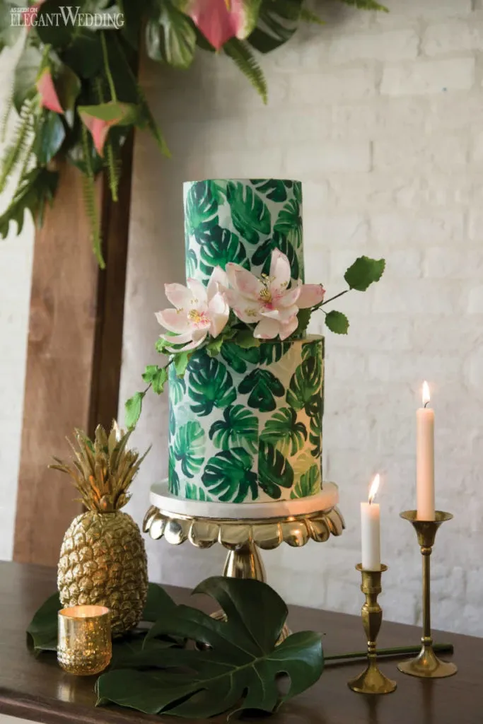 This image showcases a beautiful wedding cake by Pâtisserie Montebello, categorized under Wedding Cakes on the Mariage Elegant website in Canada. The cake features two tiers, elegantly decorated with a vibrant green botanical theme, complete with detailed leafy patterns. Adorning the cake are delicate white flowers, adding a touch of romance and freshness. The cake is presented on a classic stand, complemented by surrounding decor that includes golden candle holders with lit candles, a gold-painted pineapple, and a large tropical leaf on the table. The setting exudes a tropical and elegant ambiance, perfect for a stylish wedding celebration.