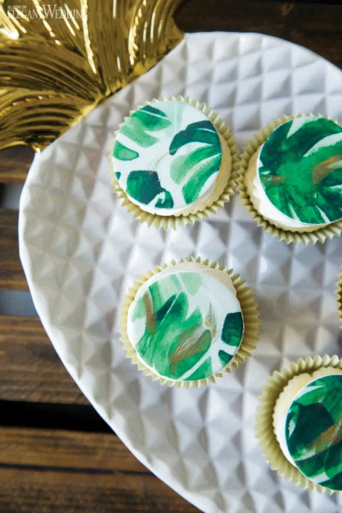 This image showcases elegant wedding cupcakes created by Pâtisserie Montebello, a vendor listed under the Wedding Cakes category on Mariage Elegant, Canada. The cupcakes are artfully arranged on a gold-accented, pineapple-shaped platter with a textured white base. Each cupcake is topped with intricate edible designs featuring green brushstroke leaves, giving a fresh and natural appearance suitable for a wedding celebration. The artistic presentation highlights the vendor's attention to detail and creativity in wedding confections.