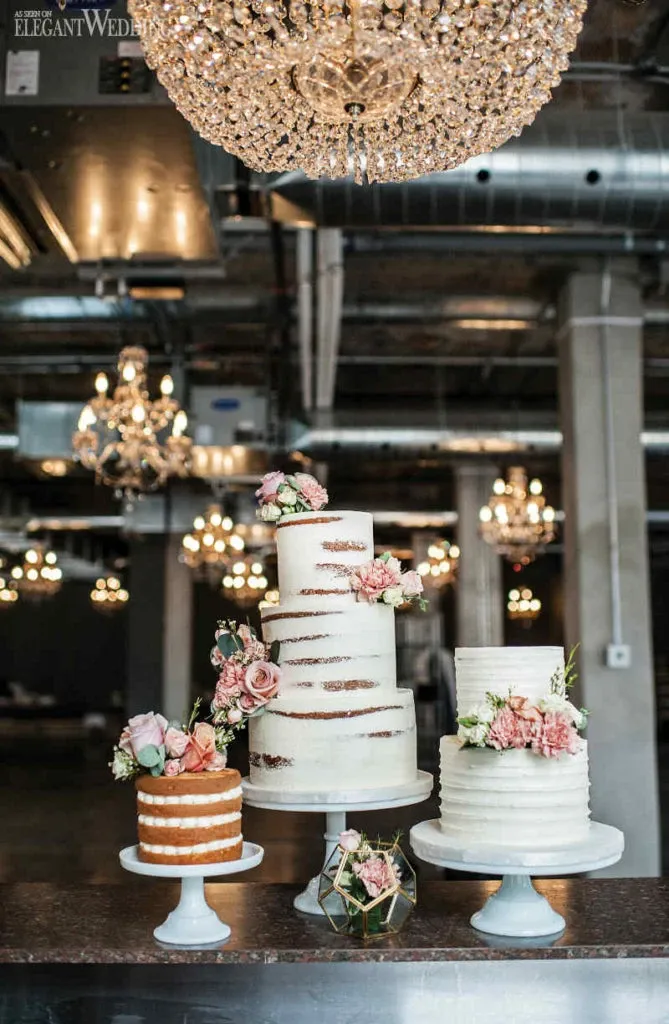 This image features a stunning display of three wedding cakes by Pâtisserie Montebello, positioned under a grand chandelier in an elegant venue. Each cake showcases unique designs suitable for weddings. The tallest cake is adorned with delicate pink and white flowers cascading down its tiered layers. The middle cake, slightly shorter, follows a textured, ribbed pattern and also boasts floral decorations, offering a classic feel. The smallest cake is a rustic naked cake with minimal frosting and topped with fresh flowers, adding a charming touch. These cakes exemplify Pâtisserie Montebello’s expertise in creating beautiful wedding cakes, perfect for adding elegance to any wedding celebration.
