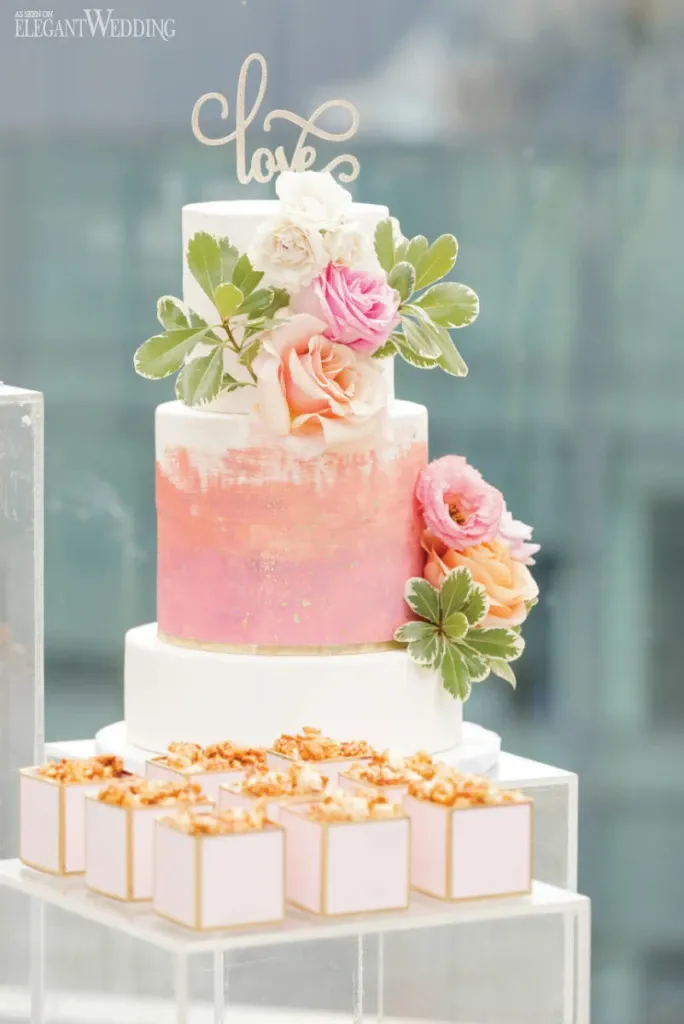 This elegant image showcases a beautifully crafted wedding cake by Pâtisserie Montebello, a vendor under the Wedding Cakes category. The cake is a three-tiered masterpiece, featuring a smooth gradient from white to a soft pink on the middle tier, adorned with delicate hand-painted textures. Fresh pink and white roses with green foliage garnish the cake, adding a touch of natural elegance. A charming 'love' topper sits gracefully atop the cake, enhancing its romantic appeal. Surrounding the cake are small, meticulously decorated dessert boxes, each with a pink and gold design, perfectly complementing the wedding theme. The setting suggests a sophisticated wedding reception with a modern and airy atmosphere.
