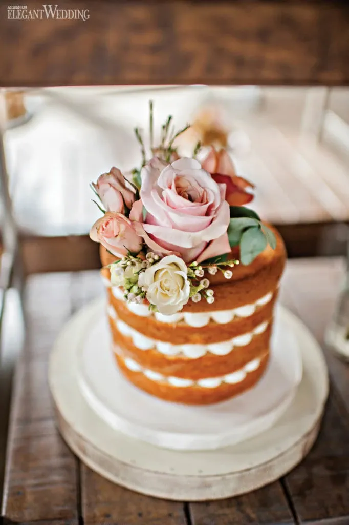 A beautifully crafted wedding cake by Pâtisserie Montebello, categorized under Wedding Cakes, is displayed. This rustic naked cake features multiple layers with smooth, creamy fillings visible between each tier. The top is elegantly adorned with a floral arrangement, including pink and white roses and delicate greenery, adding a romantic touch. The cake is set on a simple white cake board against a wooden backdrop, emphasizing its natural and elegant style, perfect for a wedding celebration.