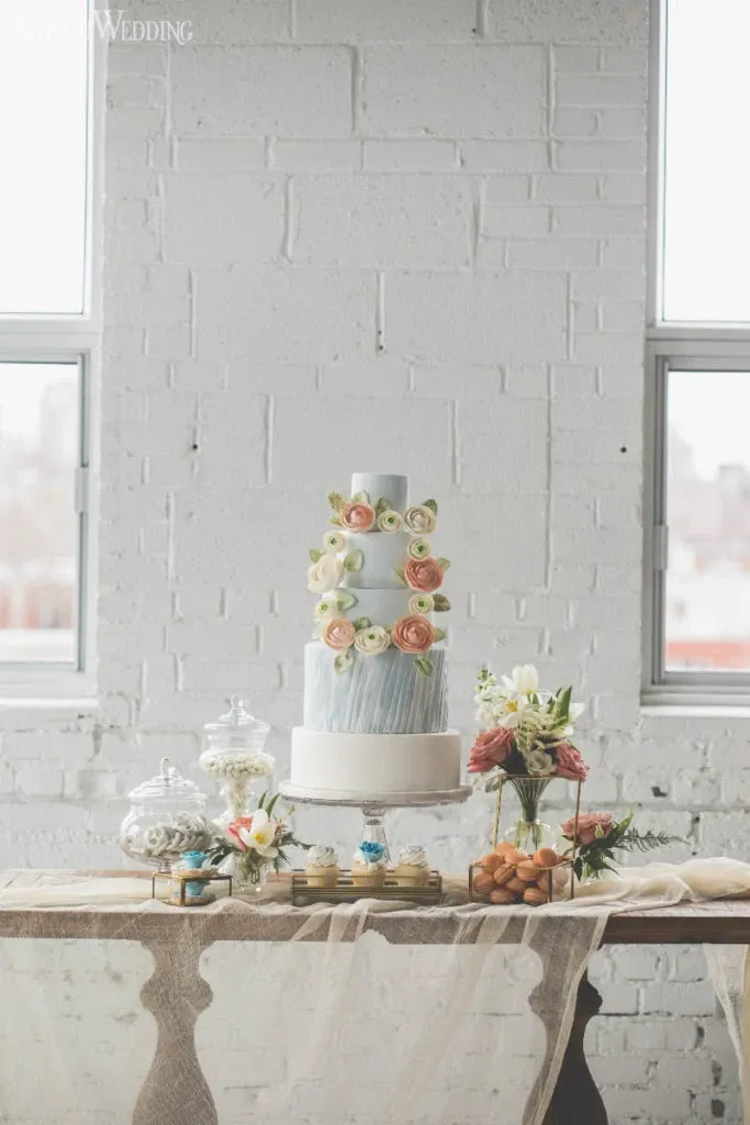 The image features a beautifully crafted wedding cake by Pâtisserie Montebello, categorized under Wedding Cakes for Mariage Elegant in Canada. This elegant cake is positioned on a decorated table against a white brick wall backdrop. The cake is multi-tiered, with a soft blue icing base adorned with delicate pink and cream floral decorations. Surrounding the cake, the table is adorned with glass jars filled with sweets and a floral arrangement, complemented by a sheer table cover that adds a touch of sophistication, perfect for a romantic wedding setting. The natural light streaming through the windows highlights the intricate details of the setup.