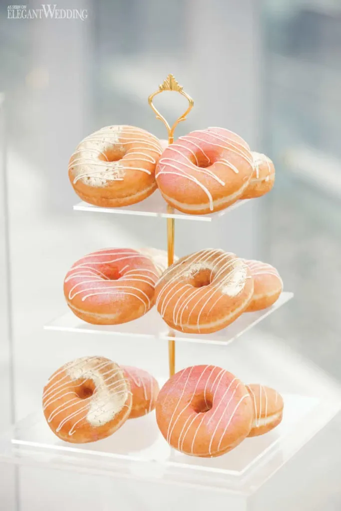 The image showcases a beautifully arranged display of wedding donuts created by Pâtisserie Montebello in the Wedding Cakes category. The donuts are glazed in a delicate pink hue and artfully drizzled with white icing, adding an elegant touch to the presentation. Displayed on a three-tiered stand with a gold handle, the setup exudes sophistication and creativity, making it a unique choice for a wedding celebration. The background features a softly blurred, bright setting that enhances the appeal of the dessert arrangement.