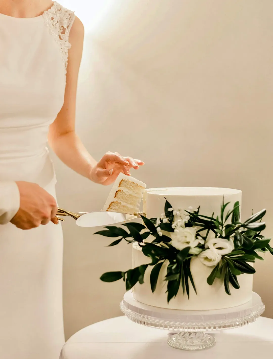 A bride in an elegant white dress holds a cake server with a slice of cake in front of a beautifully crafted wedding cake by Pâtisserie Bel Air, listed in the Wedding Cakes category. The cake features smooth white icing adorned with lush green leaves and delicate white flowers, creating a classic and sophisticated aesthetic. The cake is displayed on a clear glass stand, highlighting its detailed decorations, perfect for an elegant wedding setting in Canada.