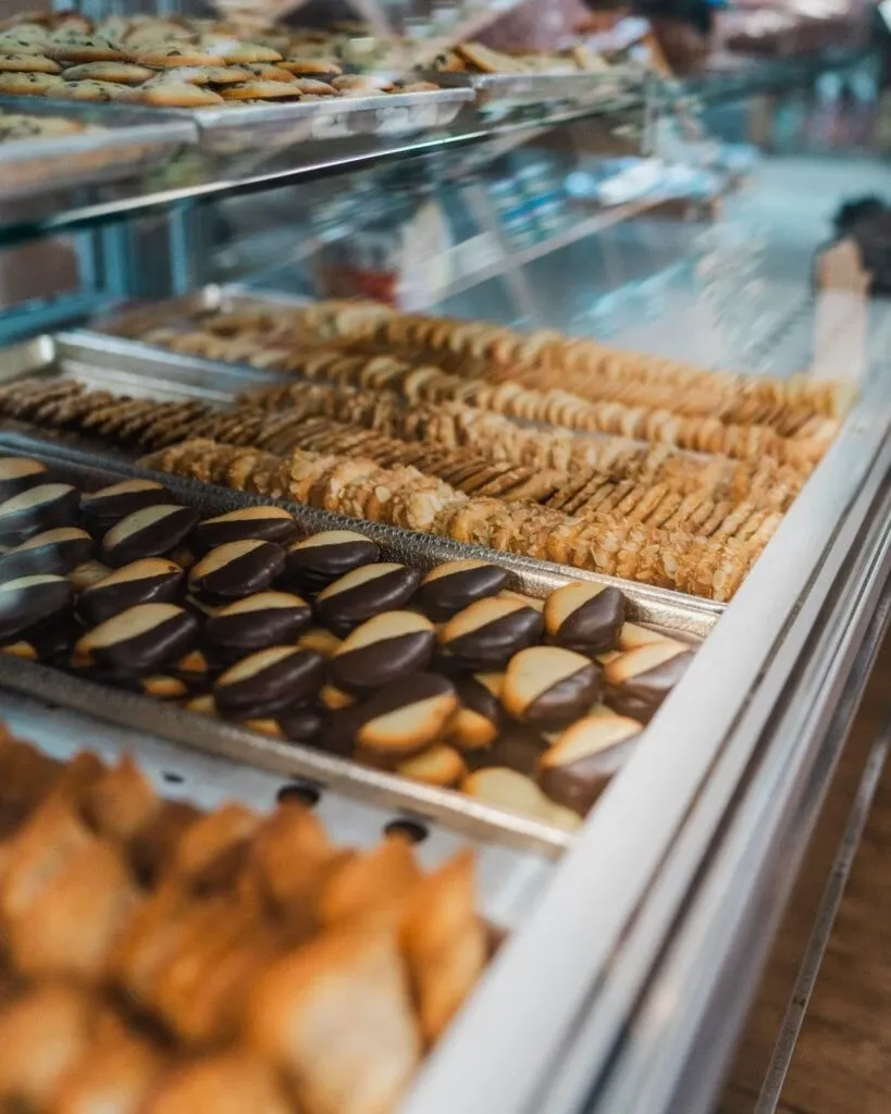 The image shows an assortment of beautifully arranged wedding pastries from Pâtisserie Bel Air, a vendor specializing in wedding cakes, displayed in a glass showcase. The foreground features rows of chocolate-dipped cookies, alongside an array of delicately baked shortbread biscuits. The backdrop reveals a variety of other pastries, highlighting the bakery's exquisite craftsmanship suitable for elegant wedding receptions. Such a display underscores Pâtisserie Bel Air's commitment to quality and presentation in the wedding category, making them a standout in the Canadian wedding planning scene.