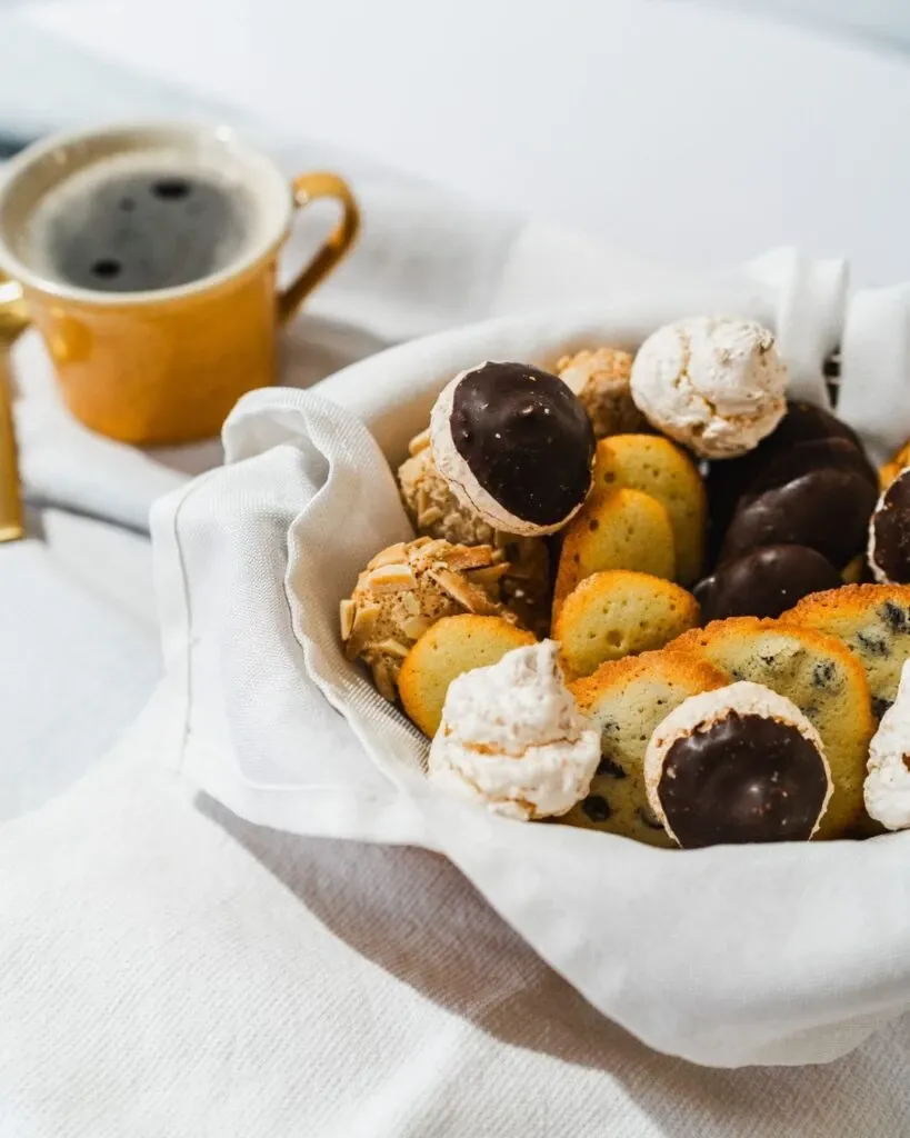 The image features an elegant display of assorted pastries by Pâtisserie Bel Air, categorized under Wedding Cakes on Mariage Elegant, Canada. A white cloth-lined basket overflows with a variety of cookies, including chocolate-dipped and almond-topped treats, alongside crispy meringue puffs. In the background, a cozy, yellow coffee cup filled with dark coffee rests on a saucer, complementing the sophisticated and inviting setting. This presentation showcases the delicate and artisanal craftsmanship ideal for wedding celebrations.
