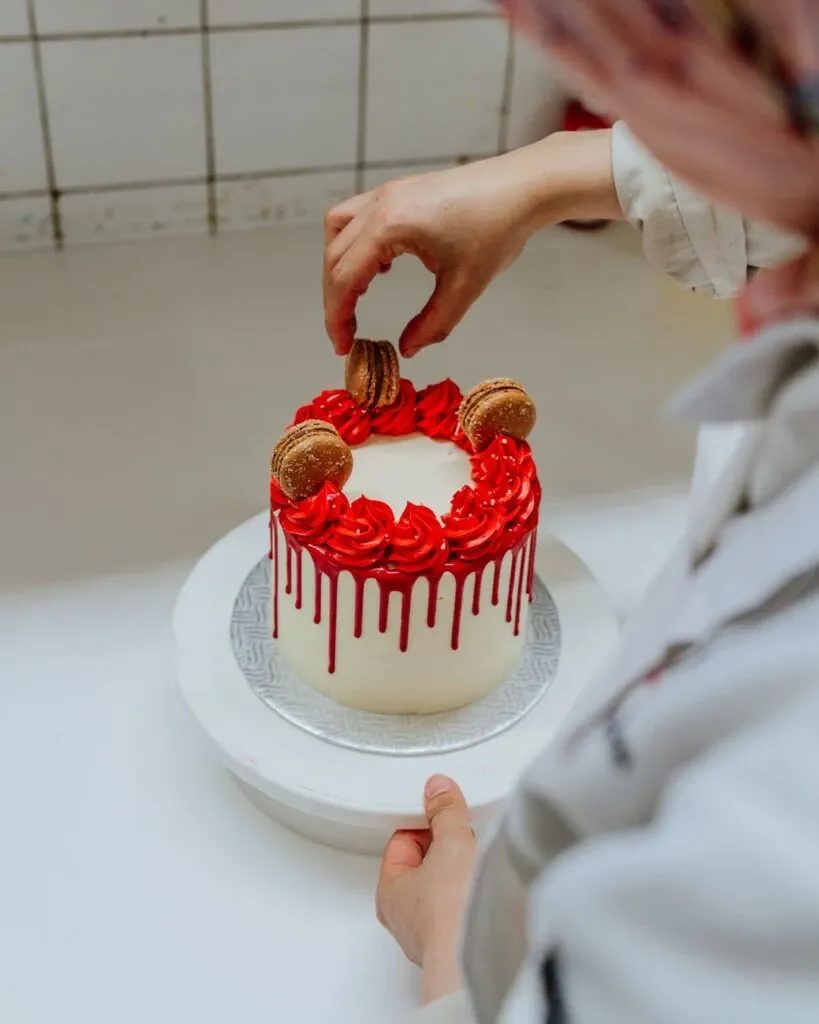 In this image for the wedding vendor Pâtisserie Bel Air, a beautifully crafted wedding cake is being delicately decorated. The cake features a smooth white frosting with elegant red drip details cascading down the sides. On top, there are red rosettes and golden macarons expertly placed, adding a touch of luxury. The hands of a pastry chef are skillfully arranging the elements in what appears to be a professional kitchen setting, underscoring the artisanal quality associated with wedding cakes in the Mariage Elegant vendor listing in Canada.