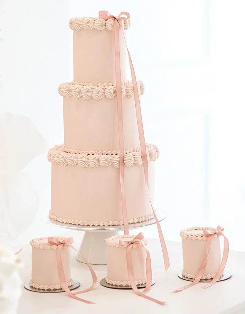 This image features an elegant three-tiered wedding cake crafted by Émotions Gâteaux, an esteemed vendor in the wedding cakes category on Mariage Elegant, Canada. The cake is meticulously decorated with a smooth pastel pink icing and adorned with delicate, scalloped icing borders on each tier. A soft pink ribbon gracefully cascades down from the top tier, adding a romantic touch. Accompanying the main cake are three coordinating mini cakes, each decorated with similar icing details and topped with matching pink ribbons, offering a harmonious and sophisticated presentation ideal for a wedding celebration.