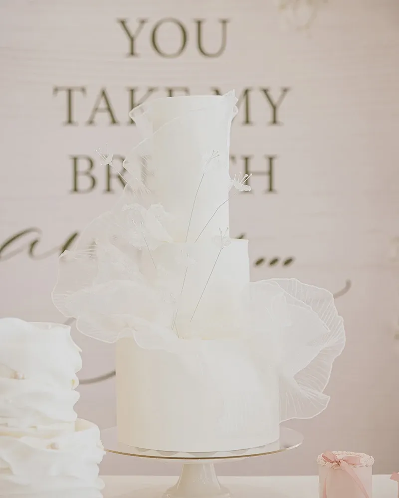 This image showcases an elegant wedding cake crafted by Émotions Gâteaux, featured in the Wedding Cakes category. The cake is a stunning multi-tiered masterpiece adorned with delicate white ruffles and intricate floral decorations, perfectly embodying sophistication and style. It stands on a simple, elegant cake stand, emphasizing its exquisite details. In the background, there is a romantic sign with partially visible text, enhancing the wedding theme. This visually striking cake is ideal for a luxurious wedding setting, highlighting Émotions Gâteaux's expertise in creating memorable wedding desserts.
