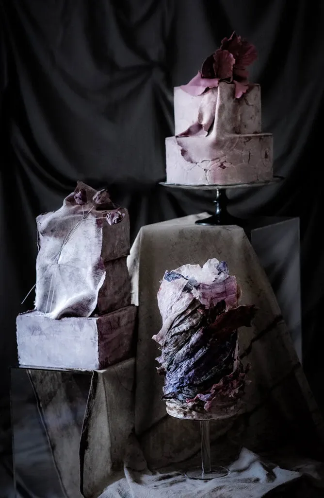 This image showcases an artistic display of wedding cakes by Émotions Gâteaux, featured in the Wedding Cakes category on Mariage Elegant, Canada. The setup includes three intricately designed cakes, each with a unique, sculptural appearance. The cakes exhibit a stunning blend of deep purple and mauve tones, with textures that resemble delicate fabric or layered petals. The centerpiece cake is elevated on a black stand, drawing attention to its elegant form and creative decoration. The background is dark, providing a dramatic contrast that highlights the cakes' vibrant colors and intricate designs, perfect for a sophisticated wedding setting.