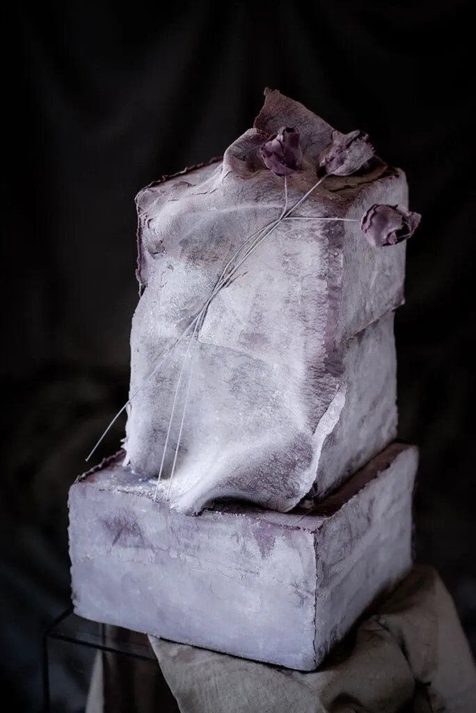This image showcases a uniquely artistic wedding cake created by Émotions Gâteaux, categorized under Wedding Cakes. The cake features a two-tier design with a rough, textured finish in muted shades of purple and gray, giving it a modern, sculptural look. Draped frosting cascades elegantly over the edges, accentuating its structured form. Delicate, dried purple flowers are carefully placed on top, adding an organic and romantic touch. The cake is set against a dark, moody backdrop, enhancing its sophisticated and whimsical aesthetic. Perfect for couples seeking a contemporary and artistic centerpiece for their wedding celebration.
