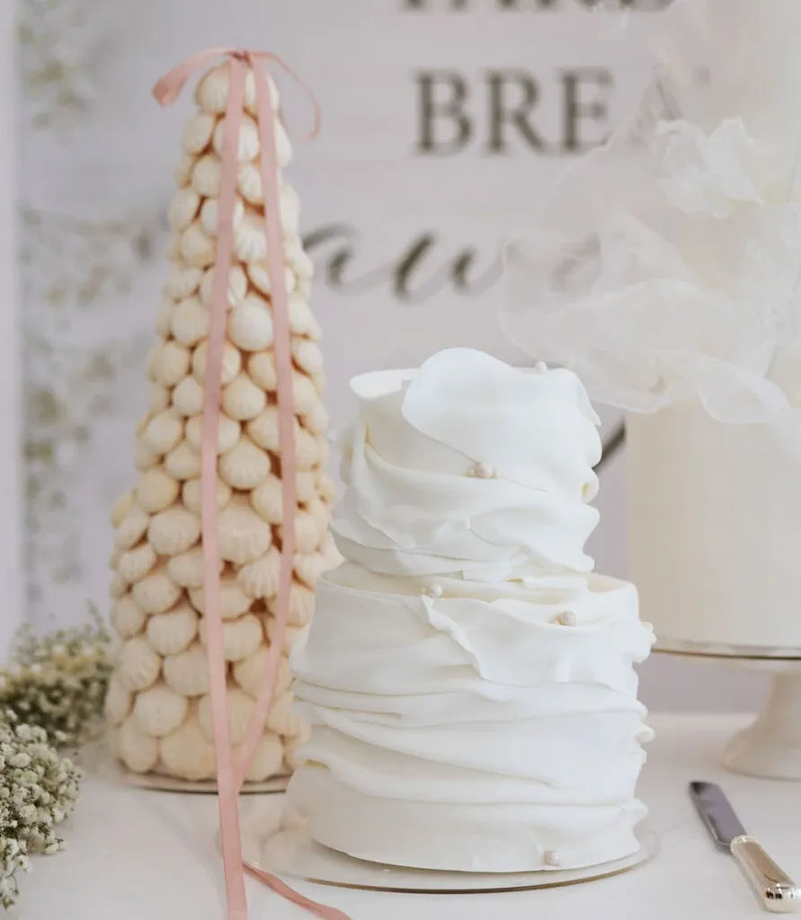 This image features an elegantly designed wedding cake by Émotions Gâteaux, showcased under the Wedding Cakes category for Mariage Elegant, Canada. The cake is a two-tiered creation adorned with white, ruffled fondant, mimicking delicate fabric folds. Beside it stands a tower of macarons tied with a soft pink ribbon, adding a touch of luxury and sophistication. Soft baby's breath flowers are arranged at the base, complementing the cake's aesthetic. The setting exudes a chic and romantic ambiance perfect for a wedding celebration.
