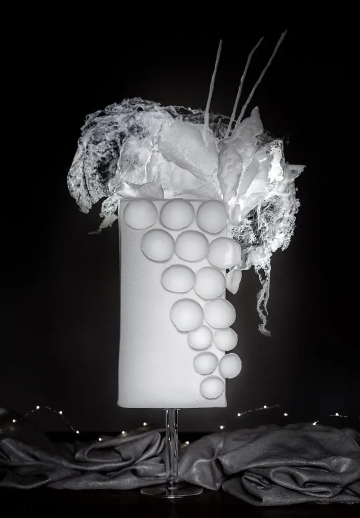 This image features a stunning wedding cake by Émotions Gâteaux, showcased in the Wedding Cakes category on Mariage Elegant, Canada. The cake is artistically designed with a sleek, tall structure and covered in smooth, white fondant. It is elegantly adorned with a unique arrangement of spherical decorations cascading down the side. At the top, there's an intricate display of delicate white sugar flowers and wispy, translucent details, adding a touch of sophistication and artistry. The cake is set against a dark background, highlighting its elegant design, making it an ideal centerpiece for a modern wedding celebration.
