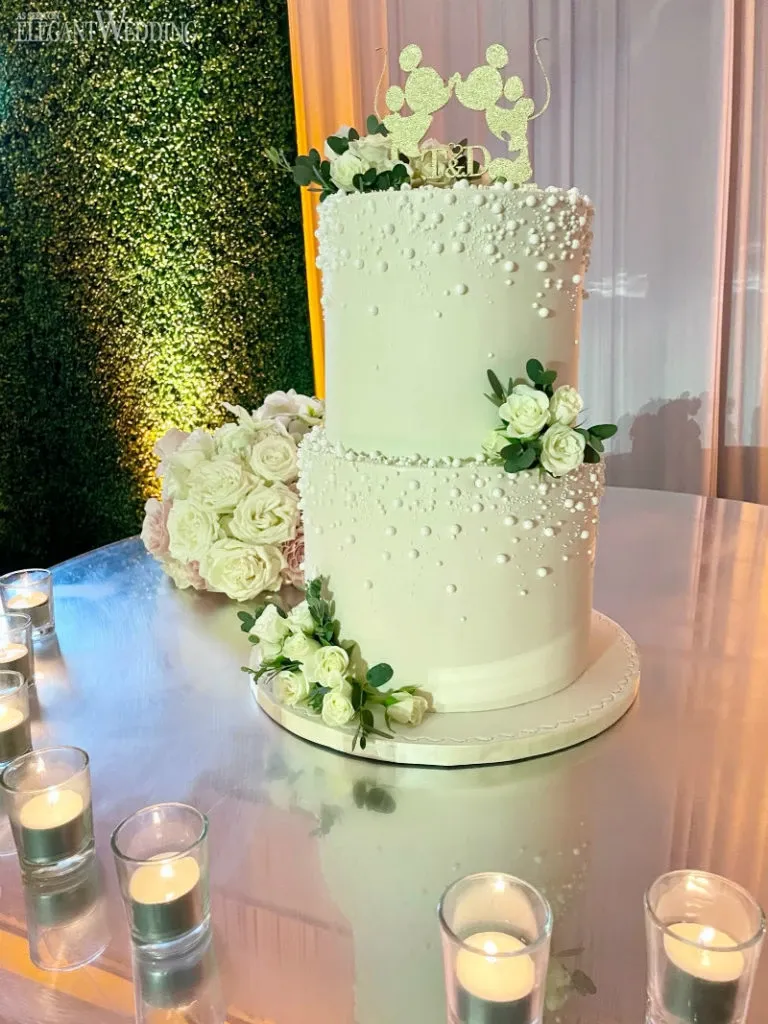 This image showcases a stunning two-tiered wedding cake crafted by Ambrosia Bakery, featured under the Wedding Cakes category for Mariage Elegant, Canada. The elegant cake is adorned with delicate white frosting and pearl-like accents, creating a classic and sophisticated look. The top is crowned with a whimsical cake topper featuring miniature mice. Soft white roses and greenery beautifully complement the cake's design, while a lush bouquet of white and blush roses is visible in the background. The cake sits on a reflective surface surrounded by small lit candles, adding a romantic ambiance perfect for a wedding celebration.