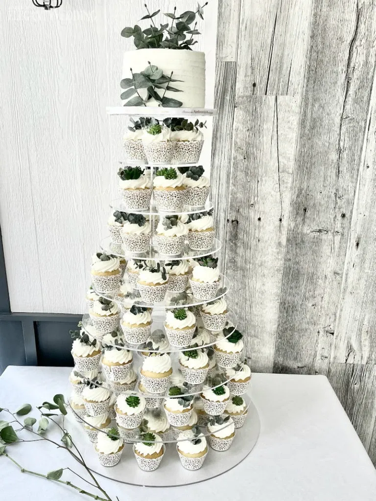 This image features a stunning display by Ambrosia Bakery, categorized under Wedding Cakes on Mariage Elegant, Canada. The multi-tiered cake stand showcases an exquisite arrangement of cupcakes elegantly decorated with white frosting and topped with delicate green foliage. At the top, a small, round cake adorns the pinnacle, accentuated by eucalyptus leaves, creating a harmonious blend of elegance and nature. The setup is situated against a rustic wooden backdrop, enhancing the overall aesthetic suitable for a refined wedding reception.