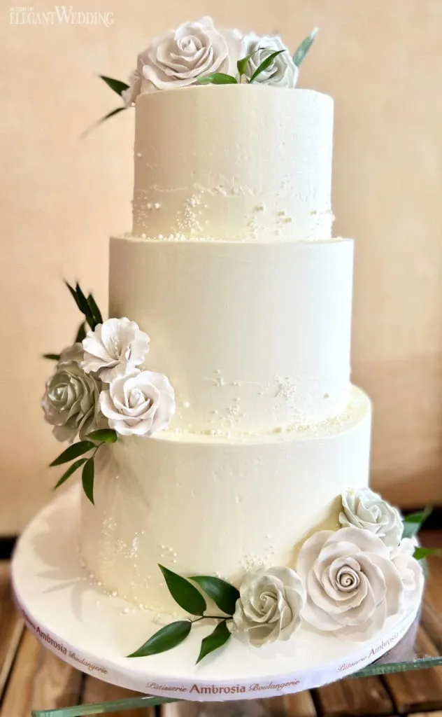 A beautifully crafted three-tier wedding cake from Ambrosia Bakery, categorized under Wedding Cakes, is displayed. The cake features smooth, white frosting with subtle textured patterns, giving it an elegant appearance. Each tier is adorned with realistic white and pale pink sugar roses, along with green leaves that add a touch of natural beauty. The cake is set on a round base, which showcases the Ambrosia Bakery branding. This exquisite creation is perfect for a sophisticated wedding celebration.