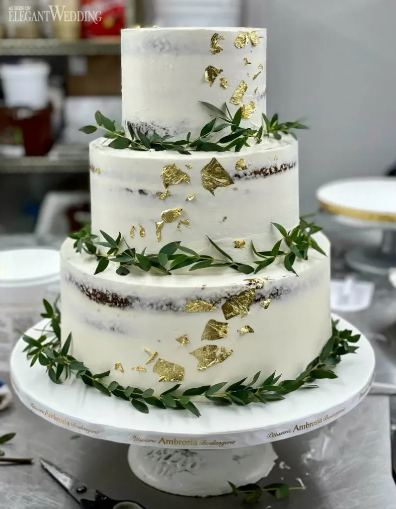 The image features a three-tier wedding cake crafted by Ambrosia Bakery, a vendor in the Wedding Cakes category for Mariage Elegant, Canada. The cake has a refined semi-naked design with smooth white frosting, elegantly accented with gold leaf embellishments. Each tier is adorned with fresh green foliage, adding a natural and elegant touch. The cake sits on a white, branded pedestal, showcasing the bakery's name, emphasizing its professional presentation in a wedding setting. In the background, a professional kitchen environment is visible, highlighting the bakery's craftsmanship.