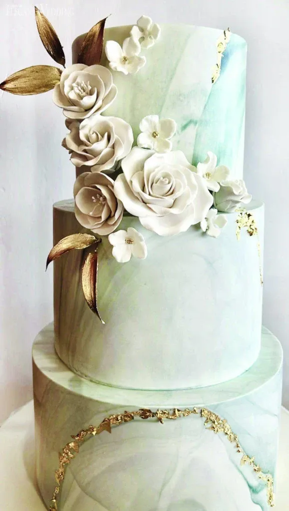 This image features a stunning wedding cake by Ambrosia Bakery, showcased in the wedding cakes category on Mariage Elegant, Canada. The cake has three tiers with a smooth, marble-like finish in soft shades of mint green and white. Each tier is adorned with delicate gold leaf accents, adding a touch of elegance and luxury. The intricate floral details, including white roses and blossoms with gold leaves, cascade gracefully down the side of the cake, exemplifying exquisite craftsmanship and elegance, perfect for a sophisticated wedding celebration.