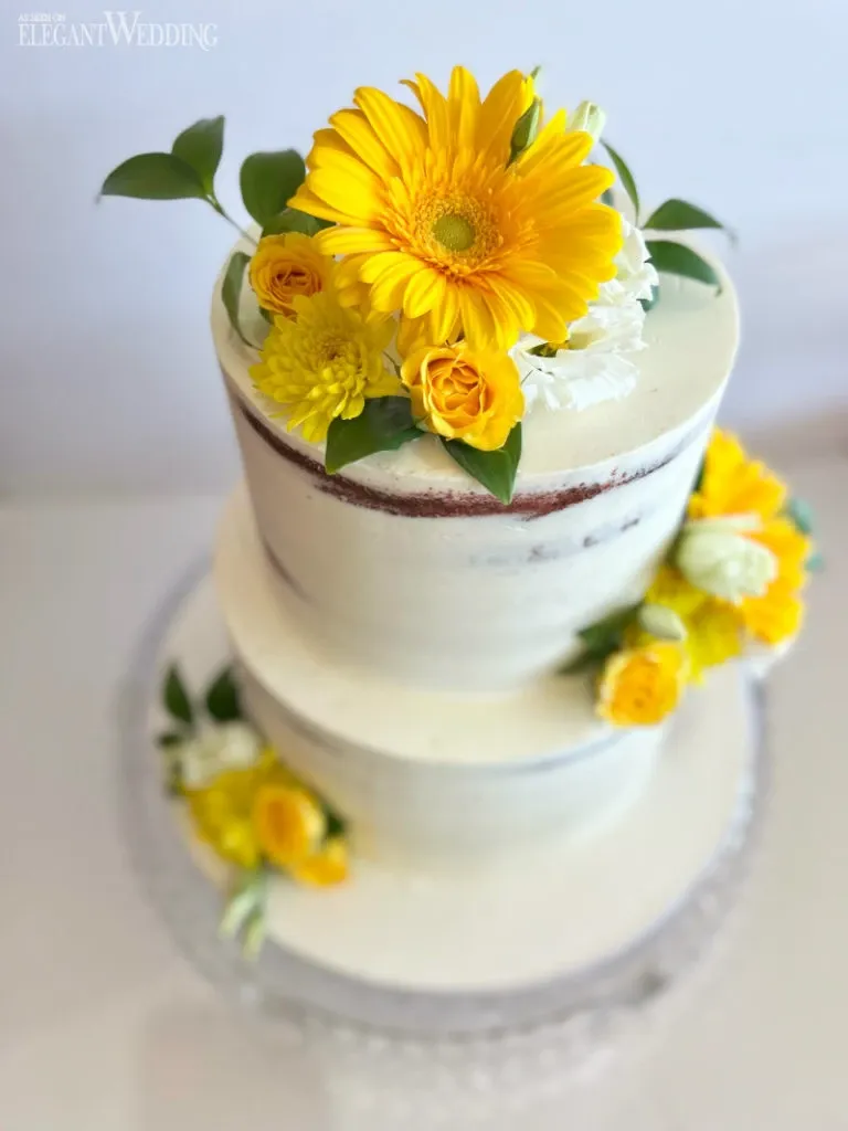 This image showcases a beautifully crafted wedding cake by Ambrosia Bakery, under the wedding category of Mariage Elegant in Canada. The two-tiered cake features a smooth white frosting with a semi-naked style, allowing glimpses of the cake beneath. It is elegantly adorned with fresh, vibrant yellow gerbera daisies and smaller yellow roses, complemented by green leaves. The cake rests on a clear, decorative glass cake stand, adding a touch of sophistication to the presentation, ideal for a stylish wedding celebration.