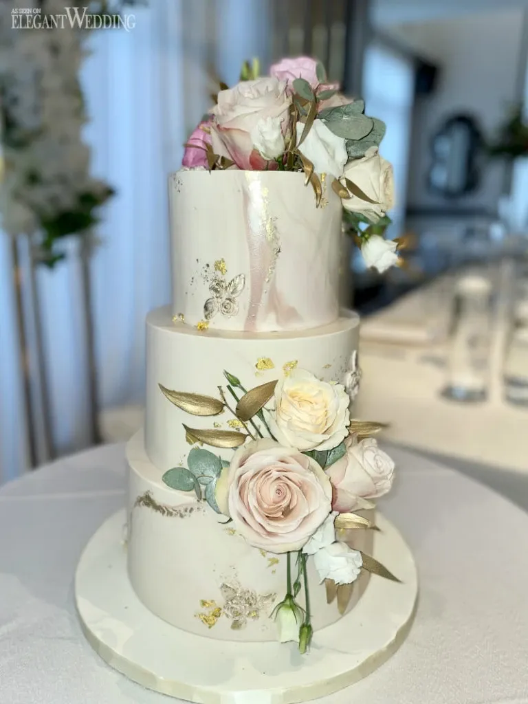 This image showcases a three-tiered wedding cake from Ambrosia Bakery, a vendor in the wedding cakes category. The cake features an elegant design with a smooth white fondant base, adorned with delicate gold leaf accents and intricate butterfly details. Each tier is beautifully decorated with an arrangement of soft pink and cream roses, accompanied by eucalyptus leaves for added greenery. The cake is set on a simple white cake stand, and the background suggests a sophisticated wedding venue setting, enhancing its romantic and luxurious appearance.