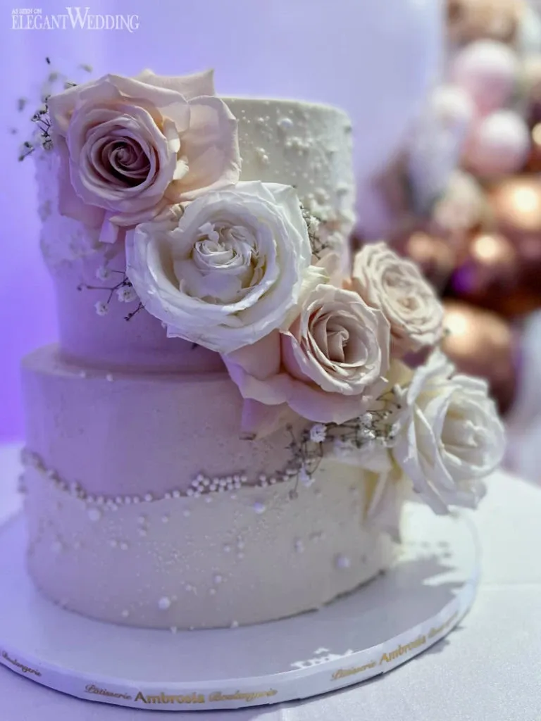This image features a beautifully crafted wedding cake by Ambrosia Bakery, falling under the wedding cakes category. The cake is elegantly designed with two tiers, each adorned with a smooth, light-colored icing. Delicate sugar pearls are sprinkled across the surface, adding a touch of sophistication. The cake is further embellished with large, realistic roses in shades of blush and white, accented by small sprigs of baby's breath. The setup captures a romantic and graceful ambiance, perfect for a wedding celebration. The elegant presentation reflects Ambrosia Bakery's dedication to artisanal quality in wedding cakes.