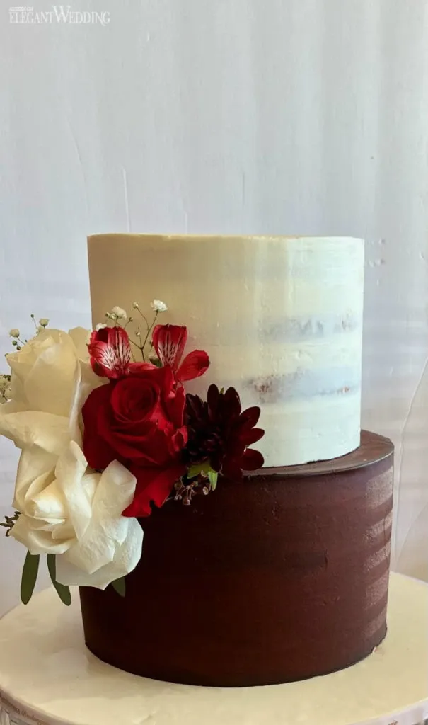 A beautifully crafted two-tier wedding cake by Ambrosia Bakery, categorized under Wedding Cakes on Mariage Elegant, Canada. The top tier features a smooth, semi-naked icing style with a subtle ombre effect, while the bottom tier is adorned with a rich, chocolate frosting. Elegant floral decorations, including red roses, a white rose, and smaller accompanying flowers, embellish the side of the cake, adding a touch of romance and sophistication to this wedding centerpiece.