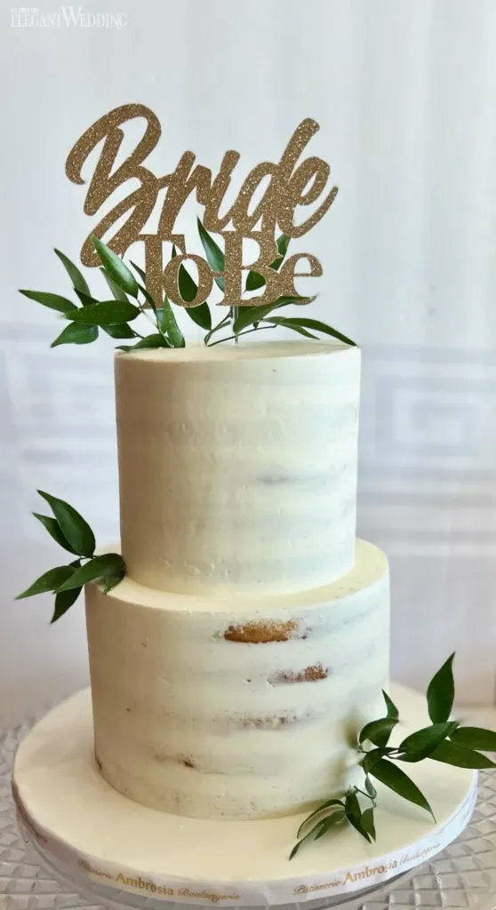 This image showcases a beautifully designed wedding cake by Ambrosia Bakery, categorized under Wedding Cakes for Mariage Elegant in Canada. The cake is a two-tier creation with a smooth, minimalist white frosting, allowing a hint of the cake's interior hue to peek through for a rustic yet elegant look. Adorned with green foliage as a natural decorative touch, the cake is topped with a glittering gold sign reading 'Bride To Be,' adding a celebratory and personal element for a bridal event. The cake is placed on a round base featuring subtle branding from Ambrosia Bakery, positioned against a soft, neutral backdrop that highlights its sophisticated design.