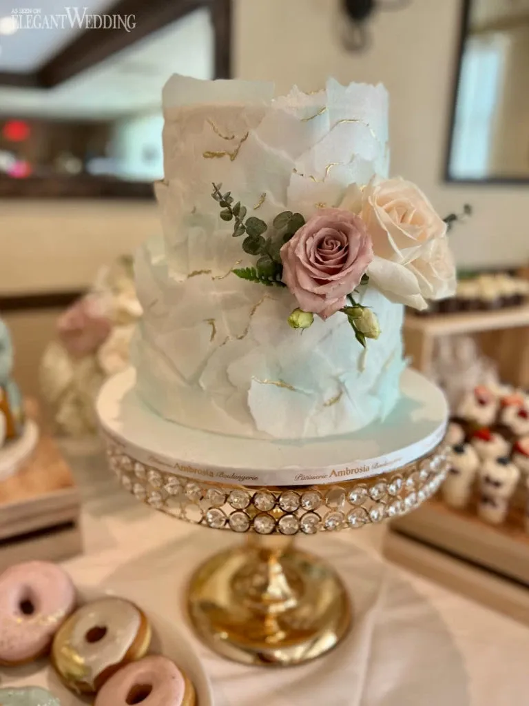 This image features a stunning wedding cake created by Ambrosia Bakery, showcased on the Mariage Elegant website in Canada under the Wedding Cakes category. The elegant two-tiered cake is adorned with delicate, textured white icing resembling floral petals, complemented by gold accents. It is beautifully decorated with fresh pink and white roses, and sprigs of greenery, adding a natural touch. The cake is displayed on a luxurious, gold stand encrusted with shimmering jewels. In the background, an assortment of other desserts, including donuts, can be seen, enhancing the celebratory atmosphere.