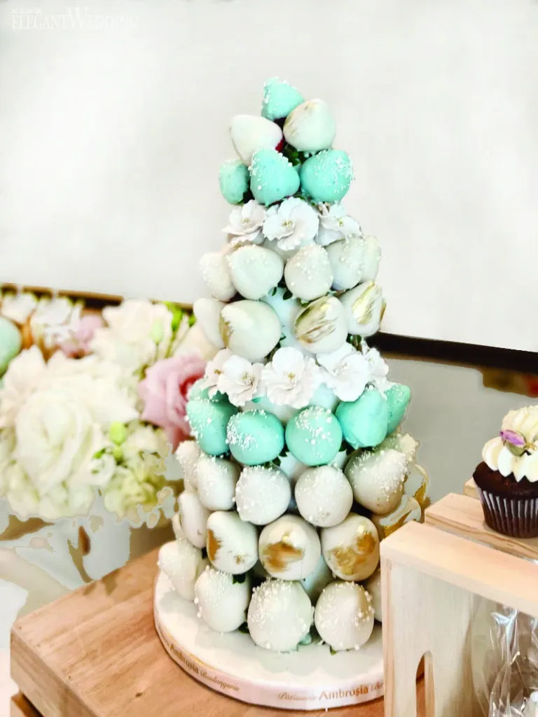 The image shows an elegant wedding cake display by Ambrosia Bakery in the category of Wedding Cakes. This unique cake is creatively designed with a pyramid of turquoise and white chocolate-covered strawberries, accented with delicate white blossoms and subtle gold brushstrokes. The cake sits on a round wooden base, adorned with the bakery's branding. In the background, a tasteful arrangement of blush pink and white flowers adds to the romantic wedding theme. A single chocolate cupcake with icing and a decorative flower is visible on the right, adding a charming touch to the overall presentation.