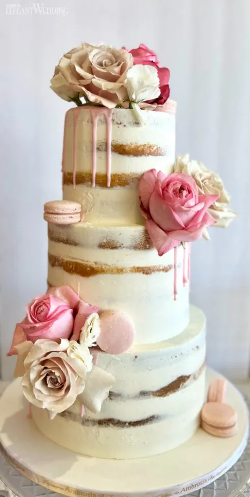 This image shows a beautifully crafted wedding cake by Ambrosia Bakery, featured in the wedding cakes category on Mariage Elegant, Canada. The cake is a three-tier semi-naked design with soft white frosting, subtly revealing the layers beneath. Adorned with elegant pink and white roses, the cake also features delicate pink macarons. The top of each tier is garnished with fresh flowers, adding a romantic touch ideal for a wedding celebration. The cake sits on a white base with Ambrosia Bakery’s name subtly printed along the edge, highlighting the vendor’s attention to detail.