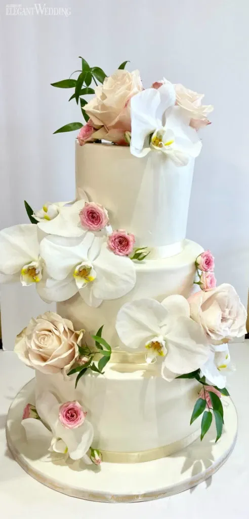 The image showcases a stunning wedding cake by Ambrosia Bakery, categorized under wedding cakes for Mariage Elegant in Canada. The cake is a three-tiered masterpiece, elegantly covered in smooth white fondant. It is adorned with a delicate arrangement of blush pink roses, small pink buds, and white orchid blossoms, adding a touch of natural beauty. Green leaves are interspersed among the flowers, providing a fresh contrast to the soft floral palette. The cake is set against a plain white backdrop, highlighting its sophisticated and classic design, perfect for an elegant wedding celebration.
