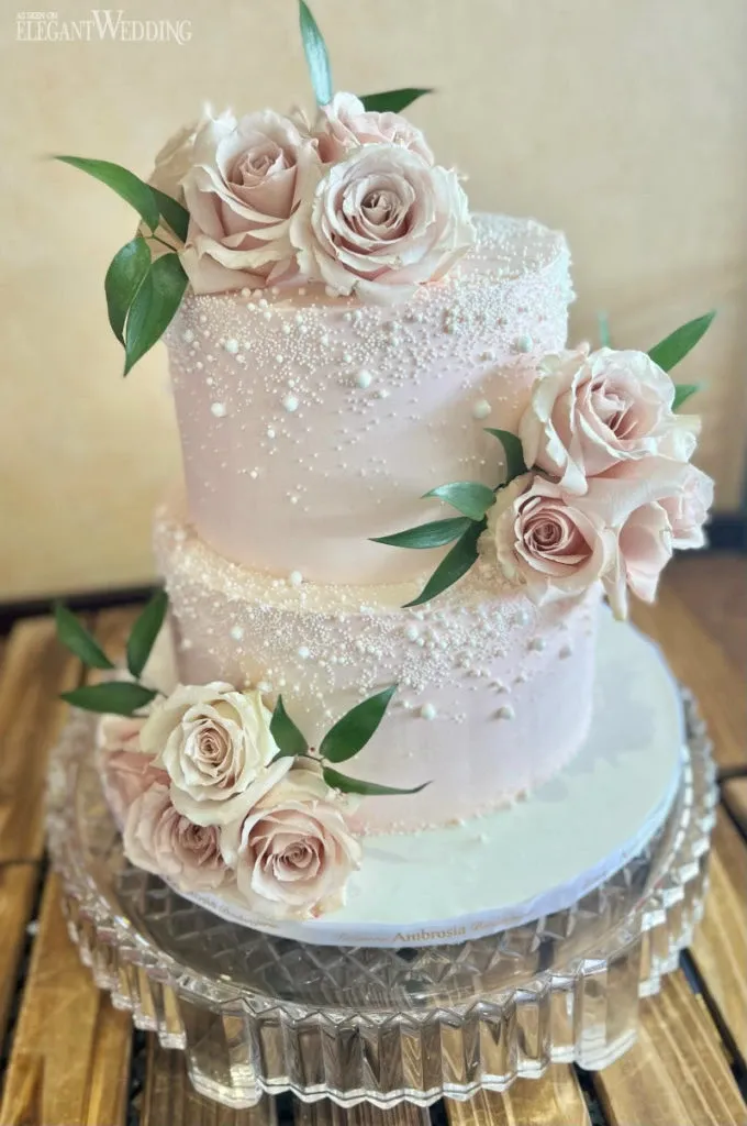 This image showcases a beautifully crafted wedding cake by Ambrosia Bakery, featured in the wedding cakes category. The cake is two-tiered, elegantly decorated in soft pink frosting. Delicate, pink roses and fresh green leaves adorn the tiers, adding a romantic touch. Intricate patterns of sugar pearls cascade down the cake, enhancing its sophisticated appearance. The cake is displayed on a clear, textured glass cake stand, emphasizing its elegant design, perfect for a wedding celebration.