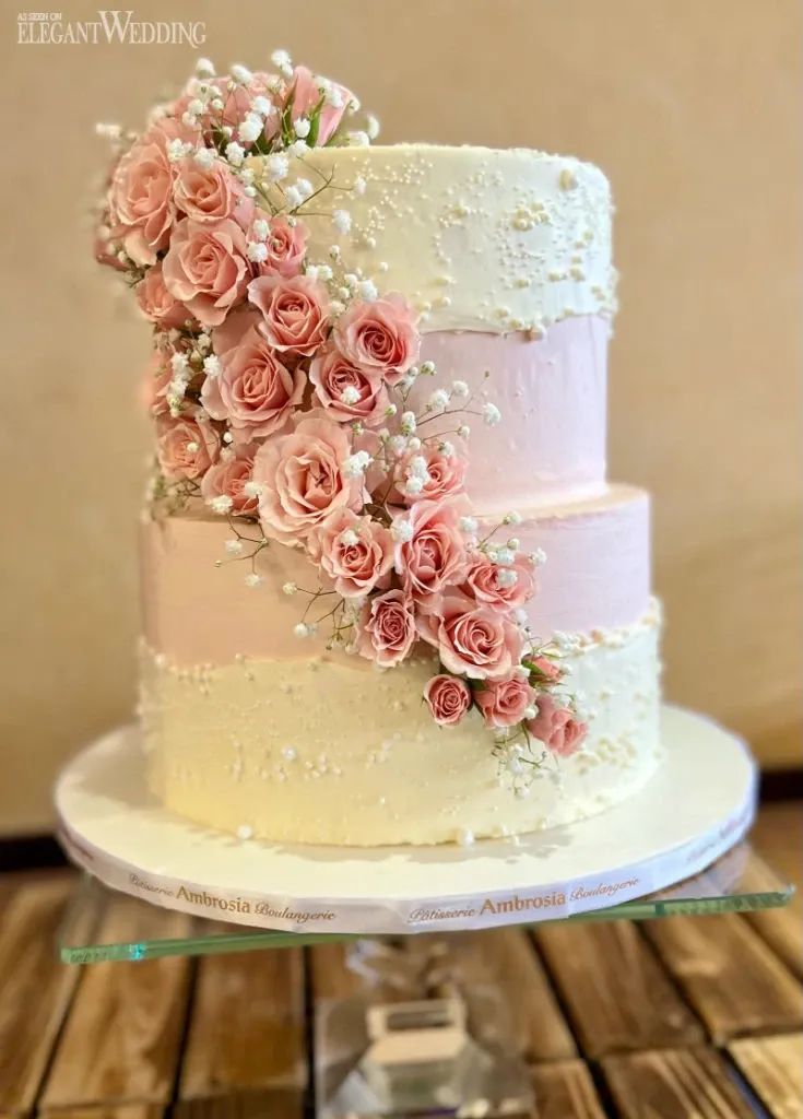 This image showcases a beautifully crafted wedding cake by Ambrosia Bakery, featured in the Wedding Cakes category on Mariage Elegant, Canada. The three-tiered cake is elegantly decorated with soft pink and cream frosting. Delicate clusters of pink roses and tiny white flowers cascade down the side, adding a touch of romantic charm. The cake sits atop a simple glass stand, emphasizing its classic and sophisticated design, perfect for a wedding celebration.