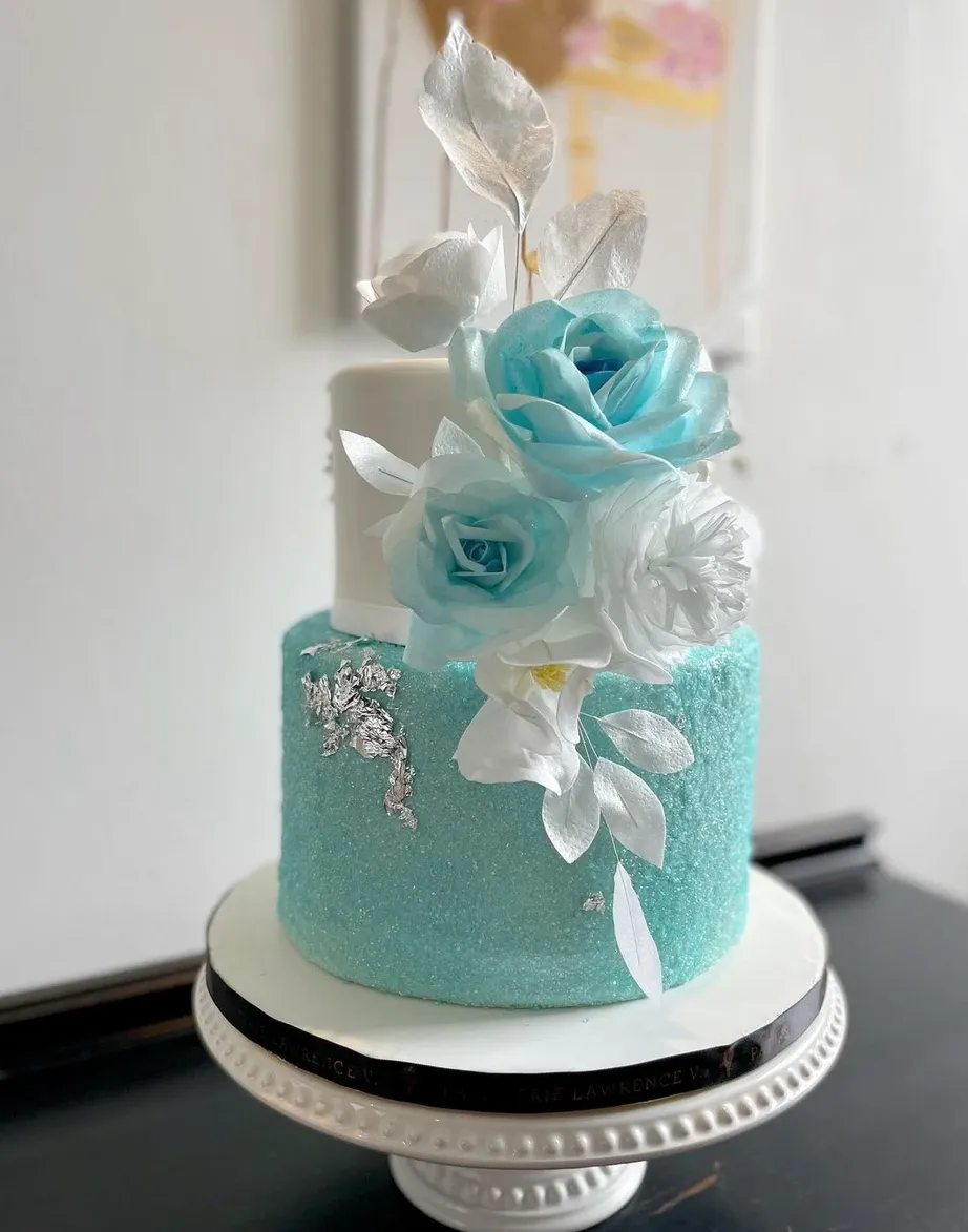 This image features a stunning two-tier wedding cake by Pâtisserie Lawrence V., displayed on a decorative white cake stand. The lower tier is covered in a textured turquoise icing, embellished with delicate silver leaf accents, while the upper tier is smooth and elegantly simple. The cake is adorned with exquisite blue and white sugar flowers and leaves, adding a touch of floral elegance. This creation perfectly represents the artistry in wedding cakes, ideal for couples seeking a modern and luxurious design.