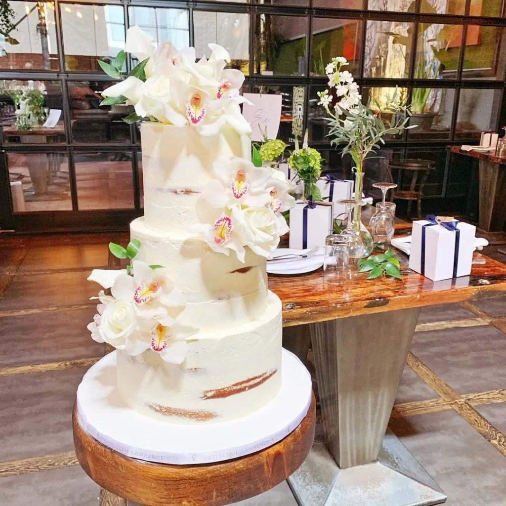 This image features a stunning three-tiered wedding cake crafted by Pâtisserie Lawrence V., a vendor listed under the Wedding Cakes category on Mariage Elegant, Canada. The cake is elegantly adorned with white and blush pink orchids and creamy roses, emphasizing a classic and sophisticated aesthetic. The cake's smooth, semi-naked icing reveals hints of the baked layers beneath, offering a modern and chic appearance. It is presented on a rustic wooden stand, situated in a warmly lit setting with flower arrangements and gift boxes in the background, evoking a sense of romantic elegance and celebration.