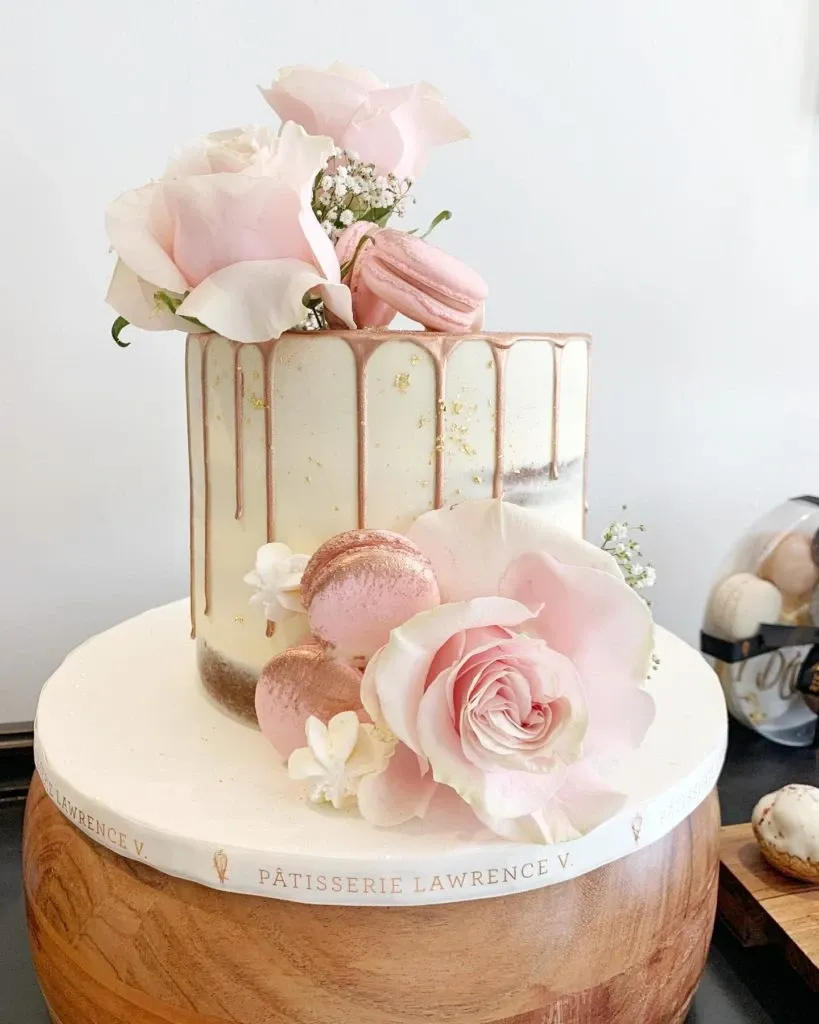 A beautifully crafted wedding cake by Pâtisserie Lawrence V., falling under the Wedding Cakes category, showcased in a minimalist style. The cake is adorned with delicate pink and white roses paired with matching pink macarons. Elegant gold accents drip from the top, complementing the pastel floral arrangement. The cake is set on a wooden pedestal with 'Pâtisserie Lawrence V.' inscribed around the base, emphasizing the vendor's brand. This elegant design is perfect for adding a touch of sophistication to a wedding celebration.