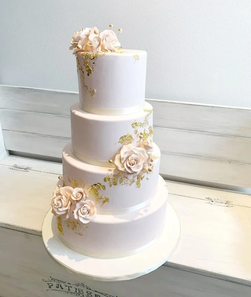 This image showcases an elegantly designed three-tier wedding cake crafted by Pâtisserie Lawrence V., a vendor in the Wedding Cakes category on Mariage Elegant, Canada. The cake features smooth, white fondant icing with delicate, pink sugar roses adorning each tier, complemented by intricate gold leaf accents. The tiers are perfectly stacked, creating a timeless and sophisticated appearance suitable for a luxurious wedding celebration. The cake is presented on a simple white cake stand against a neutral backdrop, highlighting its intricate details and artistry.