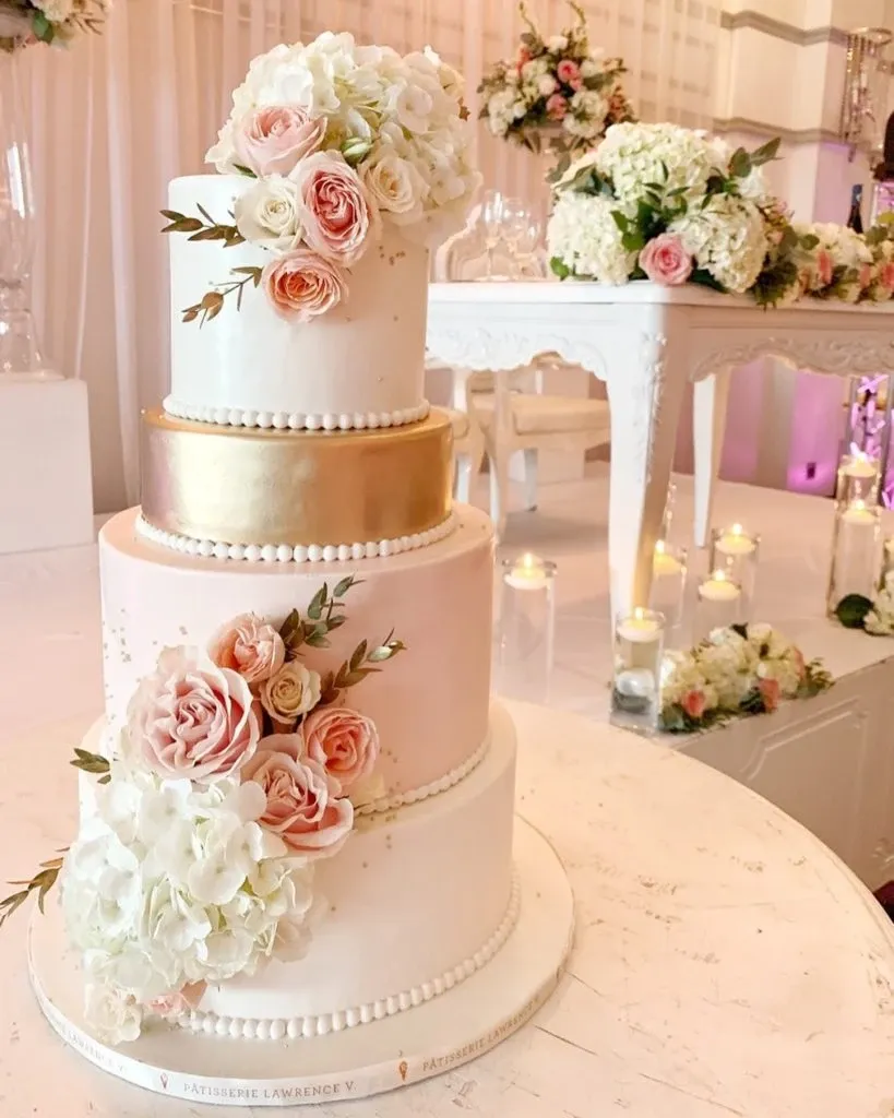 This image showcases a stunning wedding cake created by Pâtisserie Lawrence V., categorized under Wedding Cakes for Mariage Elegant in Canada. The cake is a three-tier masterpiece, elegantly decorated with soft pink and white roses, complemented by delicate greenery. The middle tier features a luxurious gold band, adding a touch of opulence to the overall design. The cake is set on a round table with a simple white cloth, surrounded by floral arrangements and soft candlelight, enhancing the romantic wedding atmosphere. In the background, a beautifully decorated table is visible, continuing the theme with matching floral decor and candles.