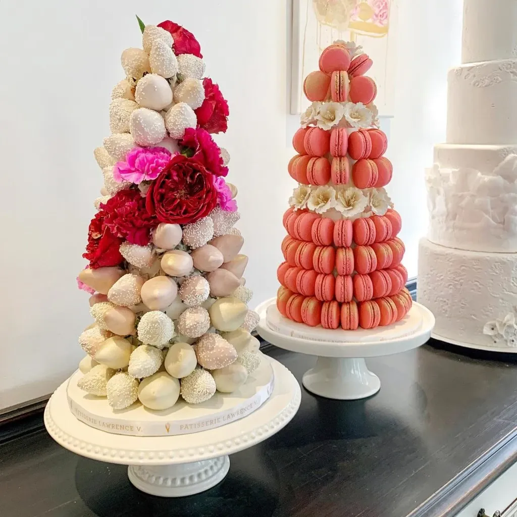 This image features two elegant wedding cake displays from Pâtisserie Lawrence V., categorized under Wedding Cakes, showcased at Mariage Elegant in Canada. The left arrangement is a striking croquembouche tower decorated with varying shades of cream and white profiteroles, interspersed with vibrant red and pink carnations, adding a touch of color and romance. The right display consists of a meticulously constructed tower of coral and pink macarons, each delicately placed on a white pedestal stand. Both creations exemplify sophisticated craftsmanship and are ideal for a luxurious wedding setting.