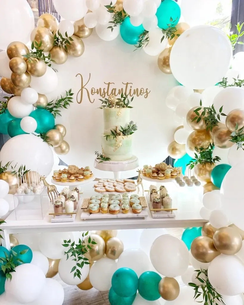This image showcases an elegant wedding cake display by Pâtisserie Lawrence V., highlighting their expertise in creating stunning wedding cakes. The centerpiece is a beautifully crafted three-tiered cake, adorned with delicate gold accents and subtle greenery, perfectly matching the refined theme. Surrounding the cake is an exquisite array of desserts, including intricately decorated cupcakes, macarons, and cake pops, all arranged on an elegant white table. Decorative balloon arches in teal, gold, and white encircle the display, adding a festive and sophisticated touch. The name 'Konstantinos' is elegantly scripted above the cake, contributing a personalized wedding element to the scene. This display exemplifies the skill and artistry offered by Pâtisserie Lawrence V. for wedding celebrations.