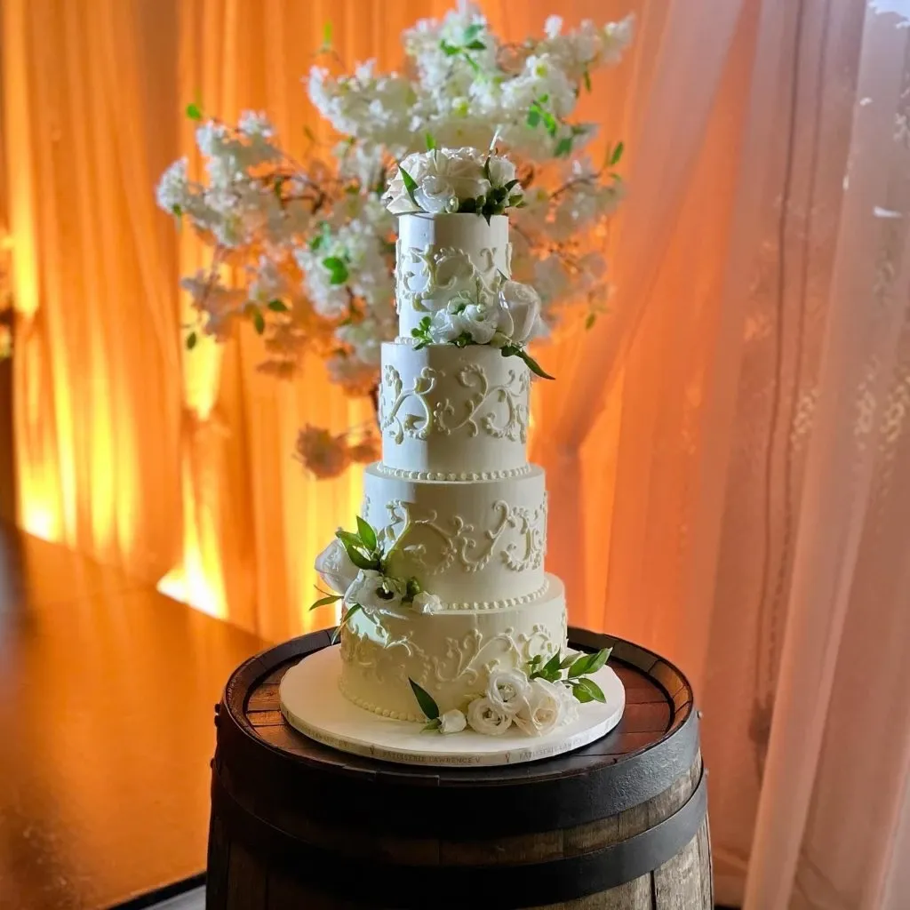 A five-tiered wedding cake created by Pâtisserie Lawrence V., part of the Wedding Cakes category on Mariage Elegant, Canada, is beautifully displayed on a rustic wooden barrel. The cake features intricate, elegant floral decorations with white roses and delicate green leaves. It is set against a warm backdrop of softly draped curtains and ethereal white blossoms, creating a romantic atmosphere perfect for a sophisticated wedding setting.