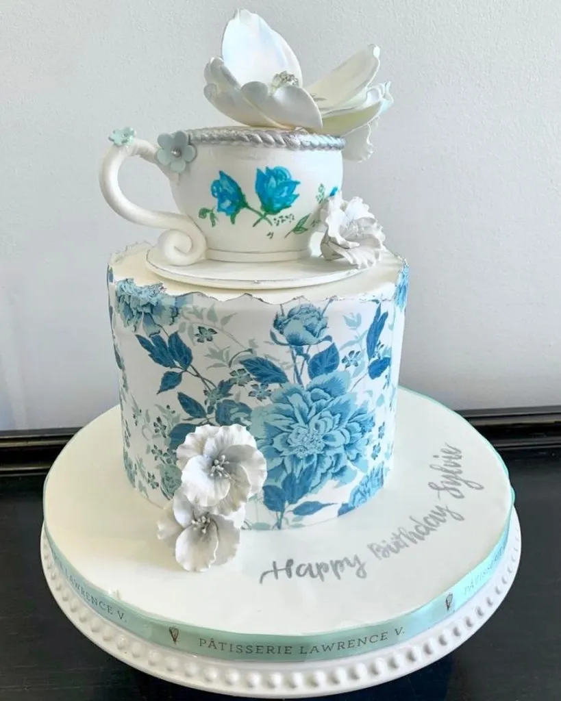This elegant wedding cake by Pâtisserie Lawrence V. showcases exquisite craftsmanship ideal for wedding celebrations. The cake features two tiers, with the top tier adorned with a delicate teacup and saucer made from fondant, elegantly decorated with a large white flower and smaller blue blossoms. The lower tier is beautifully wrapped with a detailed blue floral pattern, creating an intricate and sophisticated design. At the base, a subtle decorative band highlights the vendor's name, reinforcing its quality and reputation in wedding cakes. The cake rests gracefully on a white stand, adding a classic touch, while a personalized message, 'Happy Birthday Sylvia,' is artfully written on the cake's surface, hinting at versatile customization options for any special occasion.