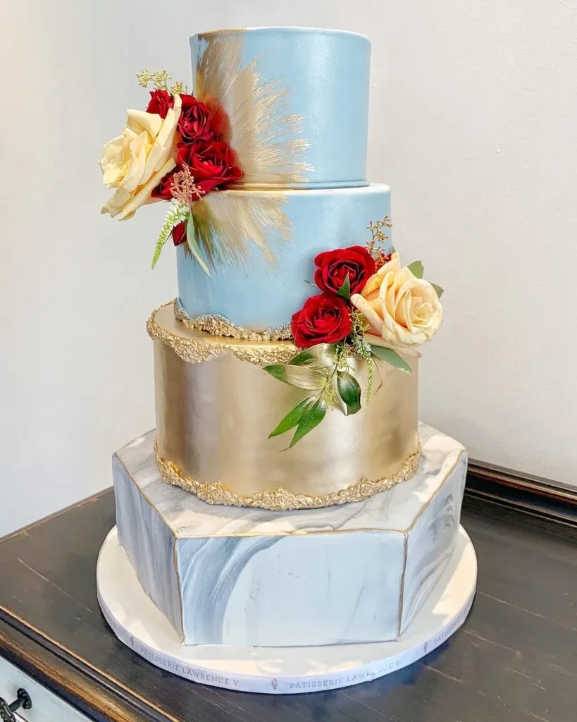 This elegant wedding cake from Pâtisserie Lawrence V., featured in the Wedding Cakes category, showcases a stunning three-tier design. The top tier is a light blue with a golden brushstroke accent, adorned with fresh red roses and soft yellow roses. The middle tier is a shimmering gold, adding a luxurious touch, while the base tier beautifully mimics a marble pattern, providing a modern and sophisticated foundation. The cake is displayed on a round cake stand, which also exhibits a marble-like appearance, enhancing its overall elegance. This artistic creation perfectly embodies the refined aesthetic of Mariage Elegant in Canada.