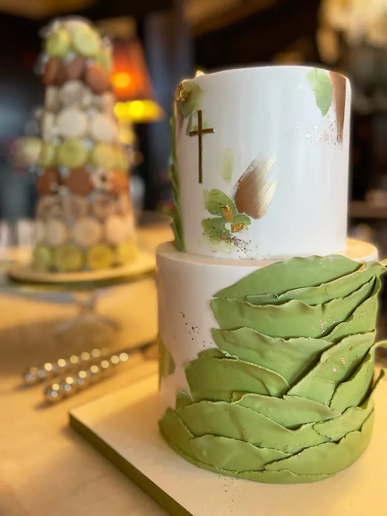 This image showcases a beautifully crafted, two-tiered wedding cake created by Pâtisserie Lawrence V. for the wedding category. The cake features an elegant white fondant base adorned with gold cross details and subtle green and gold brushstrokes on the top tier. The bottom tier is tastefully decorated with artistic green fondant leaves, adding a natural and sophisticated touch. In the background, there is a tall tower of macarons in various pastel colors, which complements the wedding cake arrangement, suggesting a combination of traditional and modern dessert offerings for weddings.