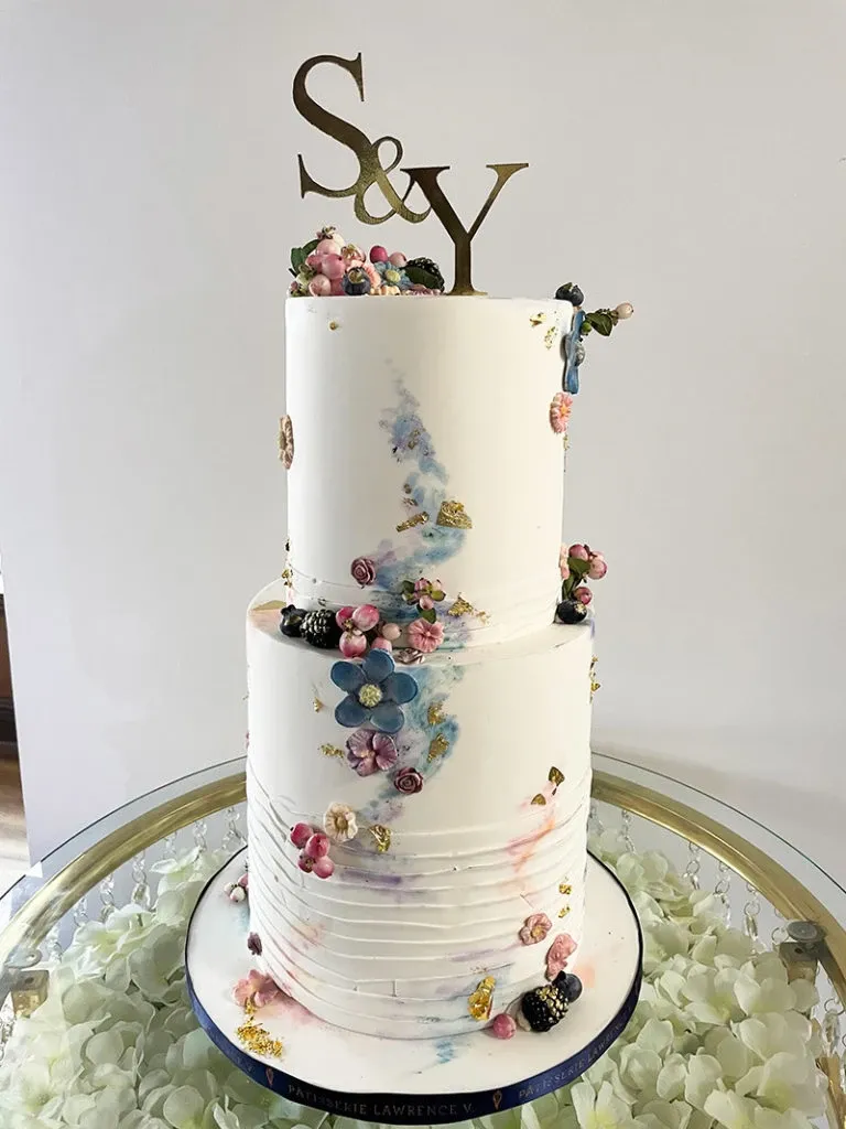 This image displays an elegant, two-tiered wedding cake expertly crafted by Pâtisserie Lawrence V., a vendor in the Wedding Cakes category. The cake features a minimalist white fondant base adorned with a colorful, artistic brushstroke design in shades of blue, pink, and gold. Delicate clusters of berries and flowers, both real and crafted, embellish the cake, adding a touch of nature-inspired beauty. The top is crowned with a monogram cake topper reading 'S & Y,' symbolizing the couple's initials. The cake is presented on a round, white floral-covered table, highlighting the sophisticated design intended for a wedding celebration by Mariage Elegant, Canada.